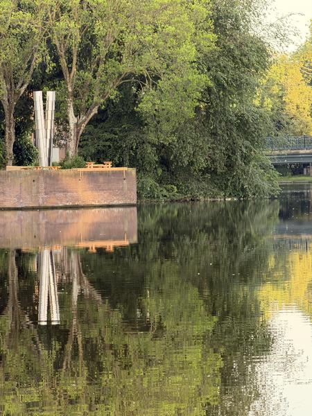 This photo captures a serene view of the Plantage Muidergracht in Plantage, Amsterdam, featuring a modern metal sculpture and wooden benches on a brick platform